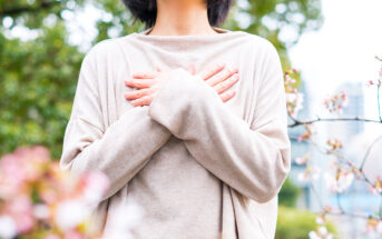 Eine Frau mit einem beigen Pullover steht in einem Garten und hat ihre Arme vor der Brust überkreuzt. Bild: Adobe Stock