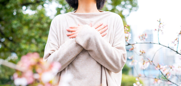 Eine Frau mit einem beigen Pullover steht in einem Garten und hat ihre Arme vor der Brust überkreuzt. Bild: Adobe Stock