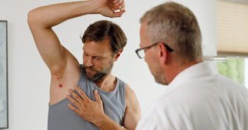 Ein Mann steht im Unterhemd vor einem Arzt und zeigt ihm rote Entzündungen in seiner rechten Achsel. Foto © Novartis