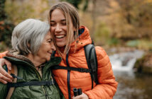 Eine erwachsene Tochter ist mit ihrer Mutter beim Wandern unterwegs. Die Tochter nimmt die Mutter liebevoll in den Arm.