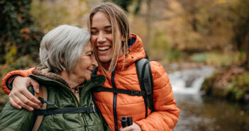 Eine erwachsene Tochter ist mit ihrer Mutter beim Wandern unterwegs. Die Tochter nimmt die Mutter liebevoll in den Arm.