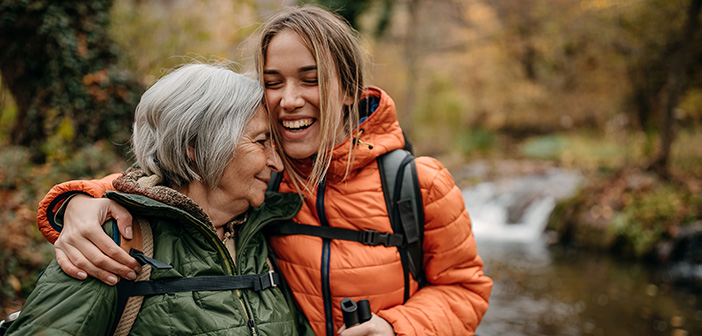 Eine erwachsene Tochter ist mit ihrer Mutter beim Wandern unterwegs. Die Tochter nimmt die Mutter liebevoll in den Arm.